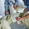 partial view of professional paramedic applying compressive bandage on leg of man near multiethnic group during first aid training seminar bleeding prevention techniques concept - Discover Tulsa Car Accident Injury Chiropractor