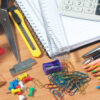 Assorted office supplies on a wooden desk including notebooks, a calculator, ruler, scissors, stapler, push pins, paper clips, pencils, eraser, and utility knife.