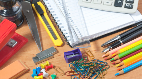 Assorted office supplies on a wooden desk including notebooks, a calculator, ruler, scissors, stapler, push pins, paper clips, pencils, eraser, and utility knife.