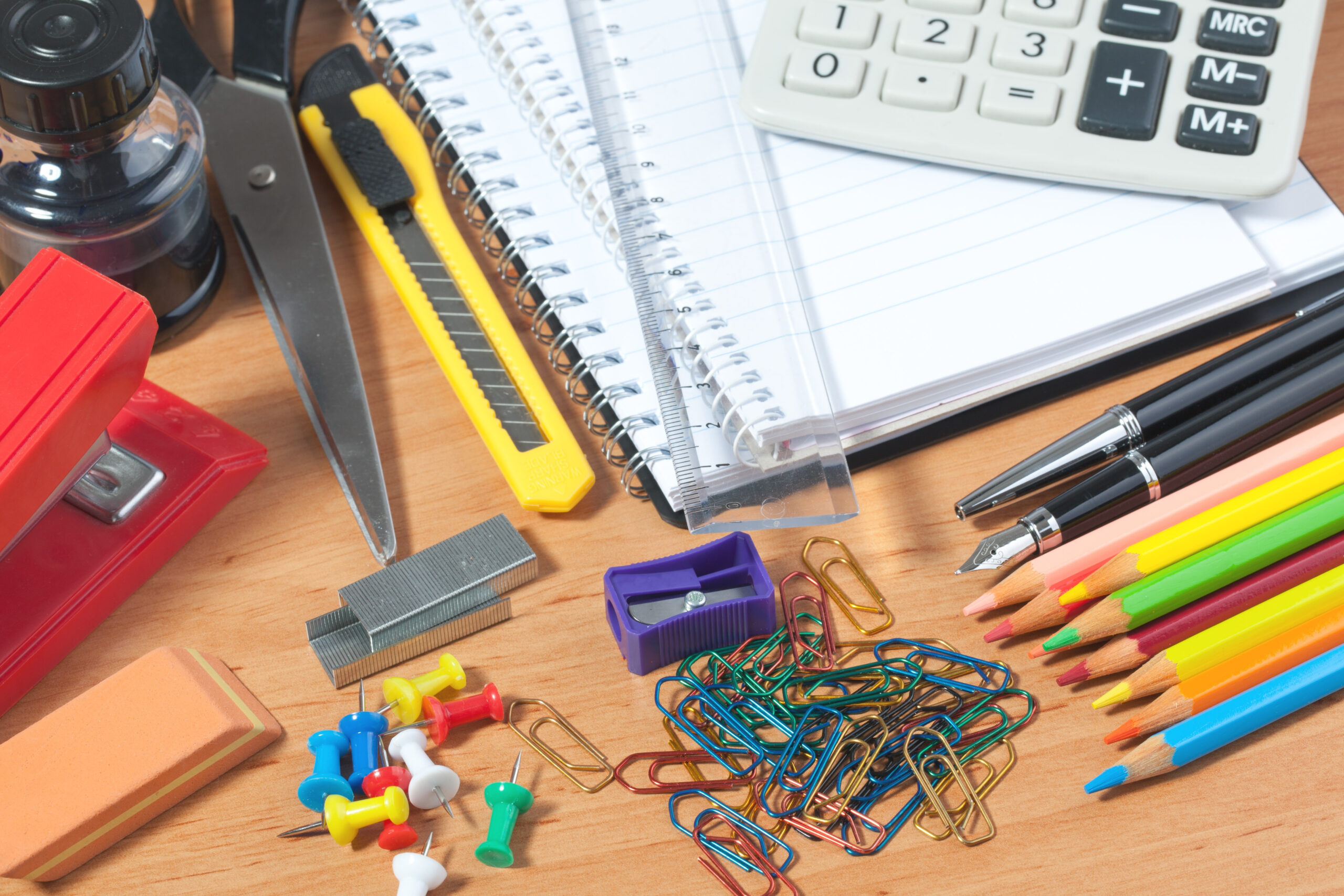 Assorted office supplies on a wooden desk including notebooks, a calculator, ruler, scissors, stapler, push pins, paper clips, pencils, eraser, and utility knife.